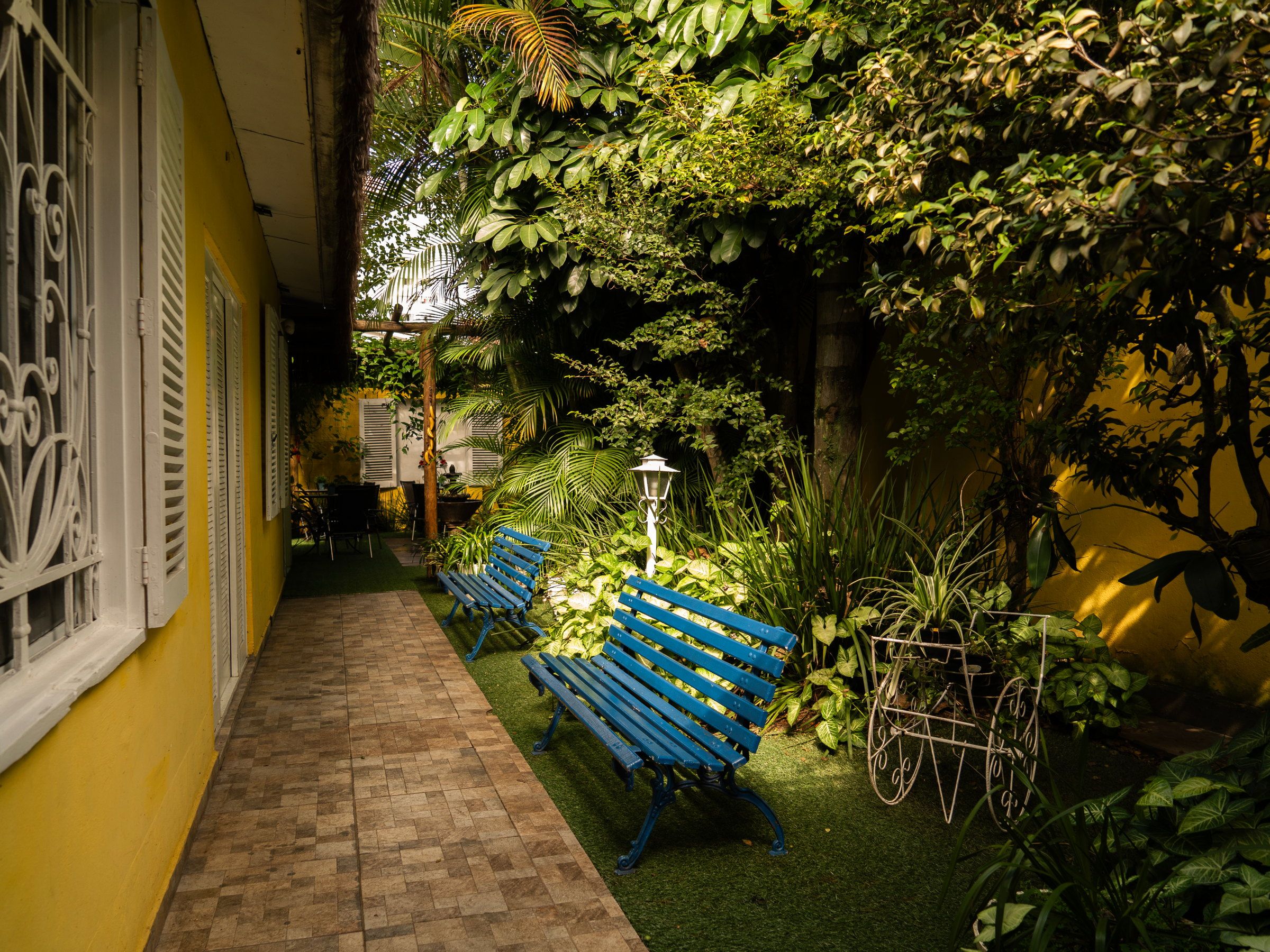 Corredor lateral da Kûara, com banco azul, plantas e janela de venezianas