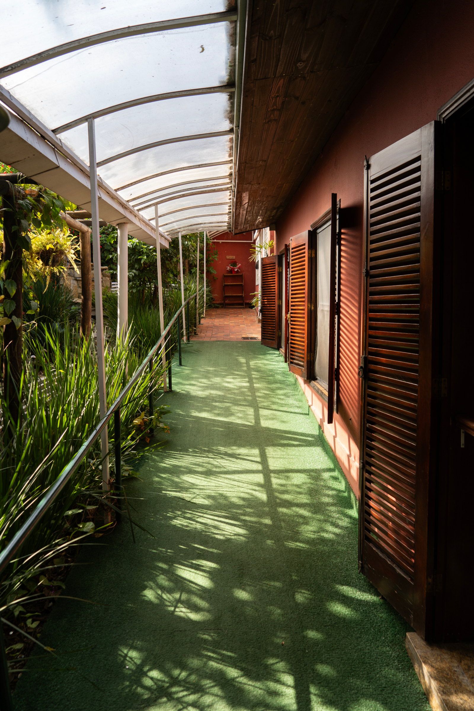 Corredor coberto ao longo da casa da Tsuru, com piso verde, venezianas de madeira e plantas