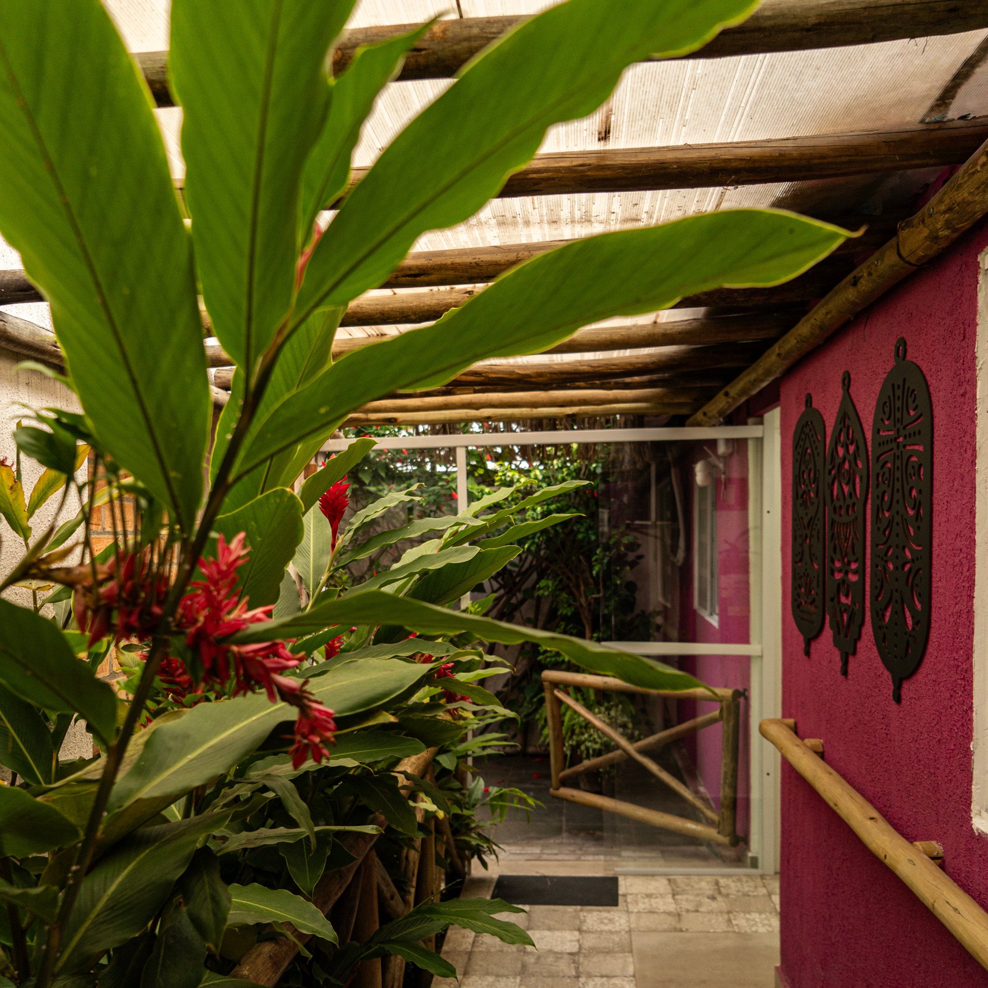 Folhagem com flores vermelhas no corredor coberto da La Vie, junto à parede magenta com enfeites de madeira e ao corrimão