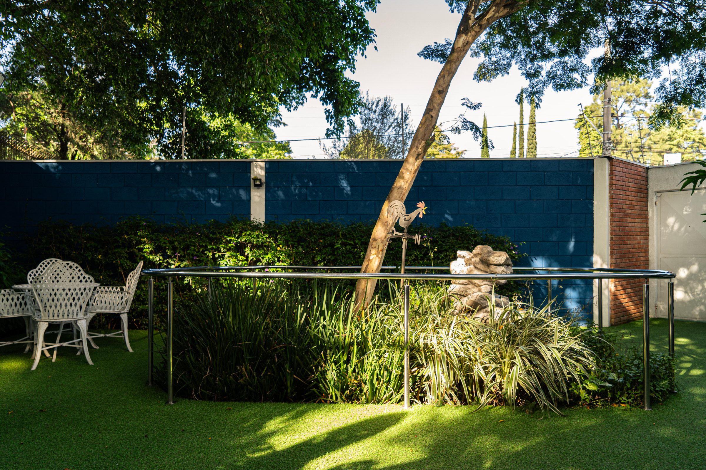Jardim do Residencial Espaço Saúde com mesa e cadeiras brancas, canteiro cercado por corrimão e uma árvore diante do muro azul