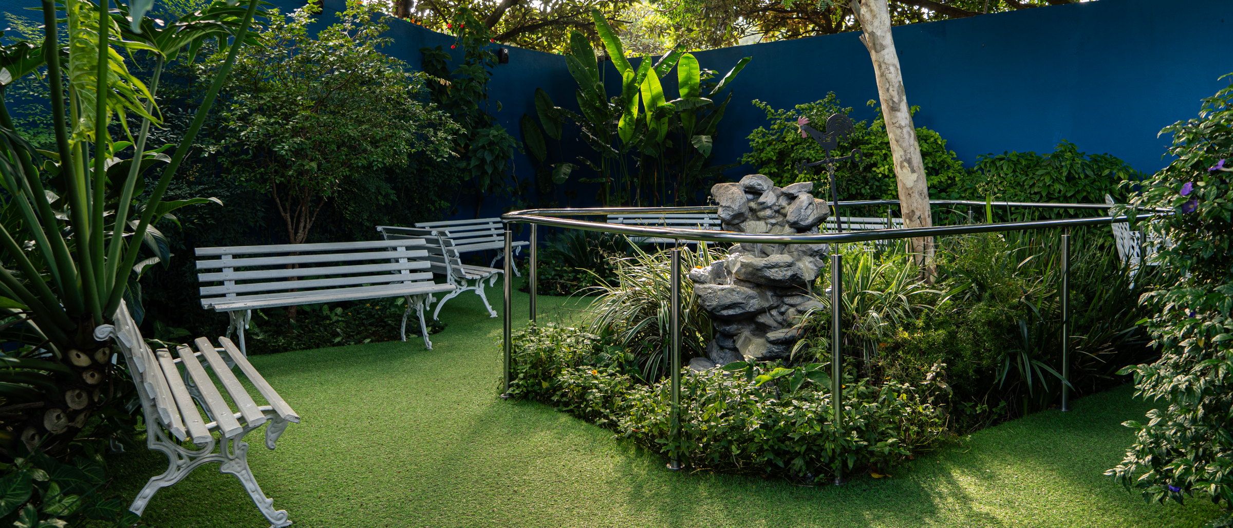 Jardim do Residencial Espaço Saúde com bancos brancos, canteiro com fonte de pedra, árvores e o muro azul