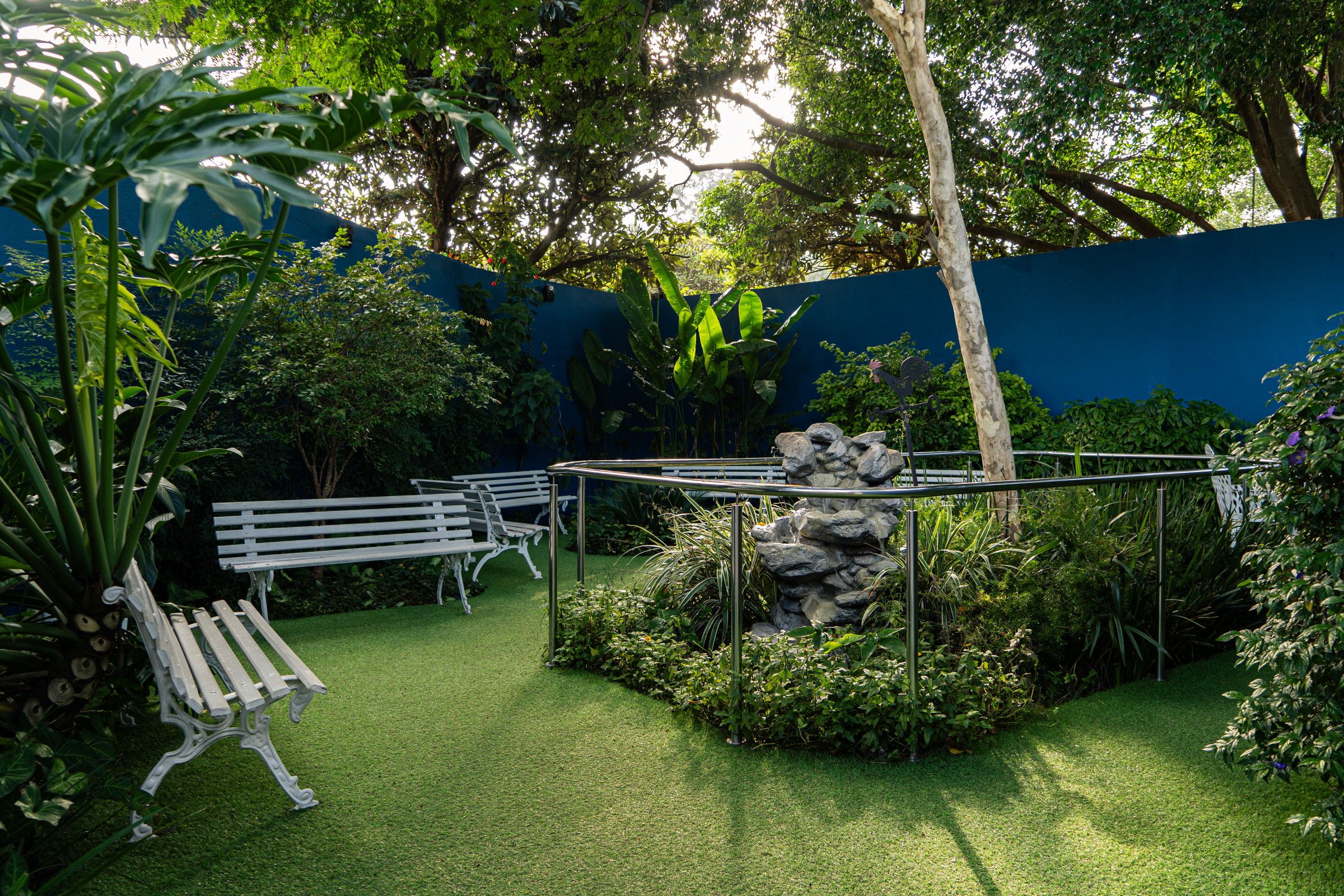Jardim do Residencial Espaço Saúde com bancos brancos, canteiro com fonte de pedra, árvores e o muro azul