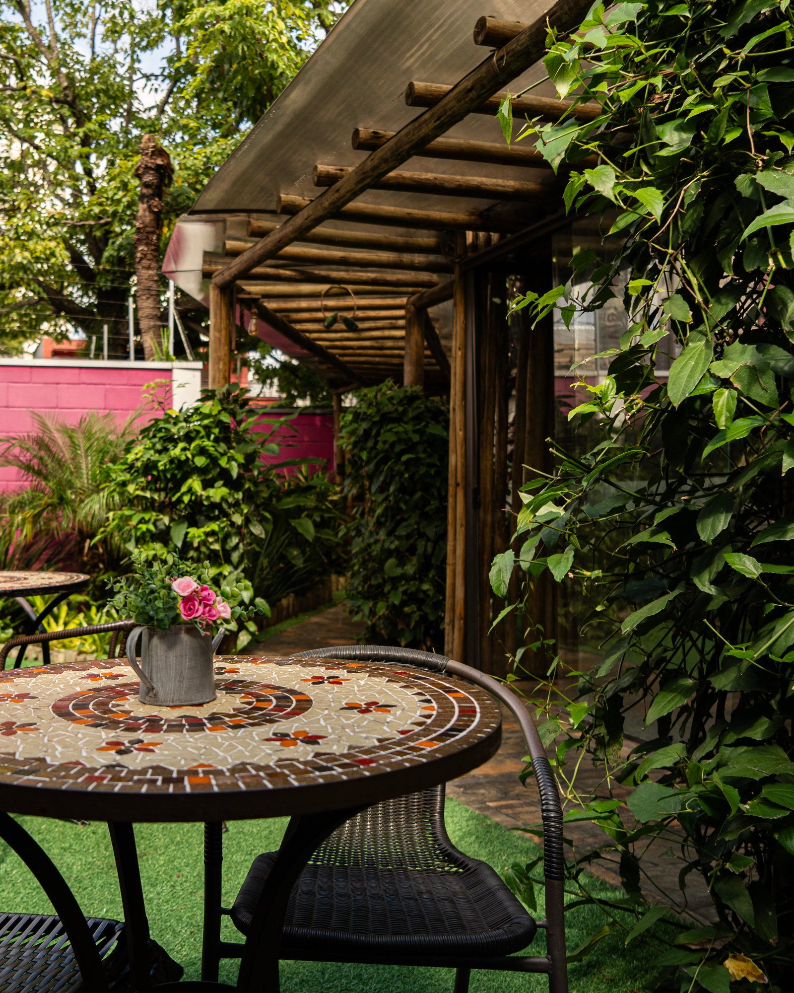 Mesa de jardim com vaso de flores sob o pergolado da La Vie, com a parede magenta ao fundo