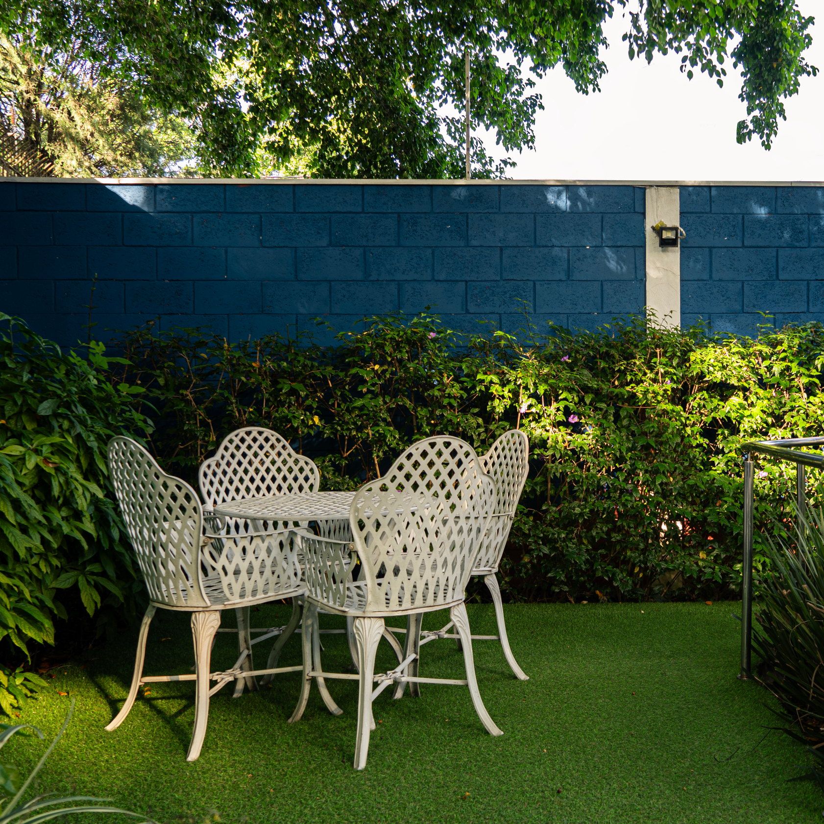 Mesa e cadeiras brancas de ferro rendado no jardim do Residencial Espaço Saúde, junto à cerca viva e ao muro azul