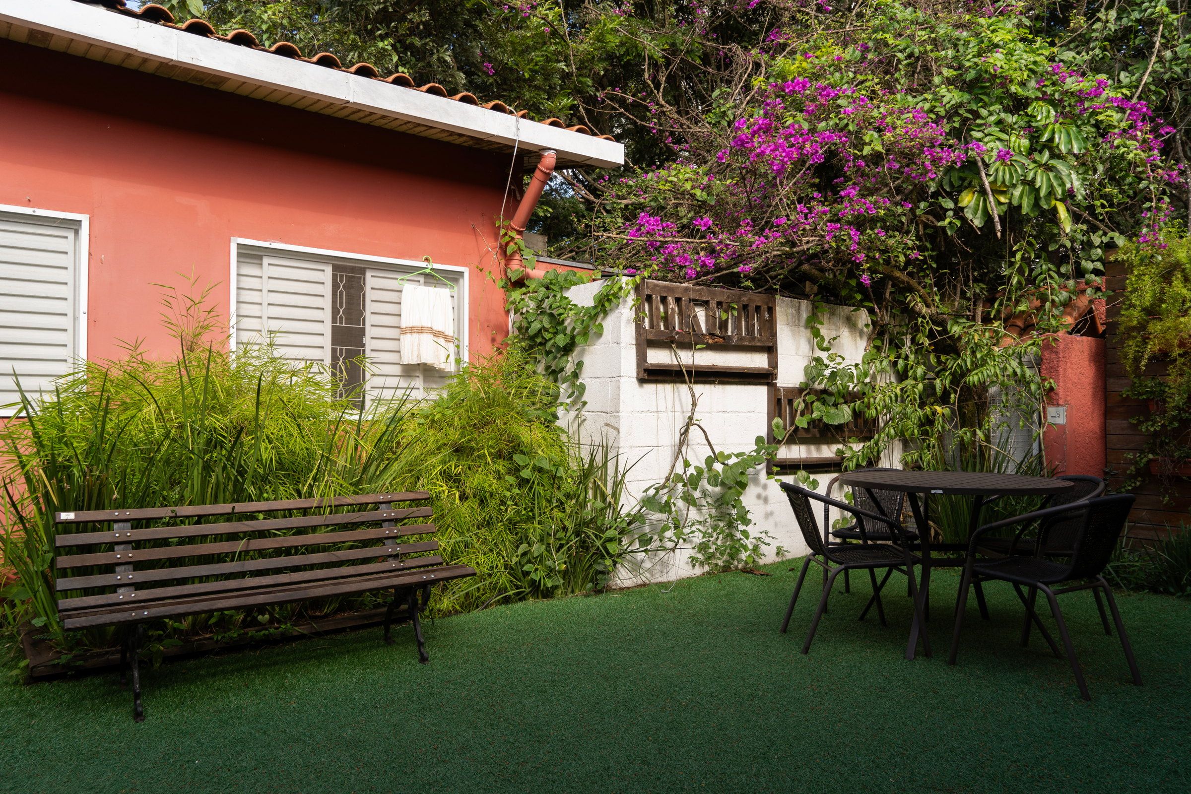 Pátio da Tsuru com banco de madeira, mesa e cadeiras, junto à casa terracota e a uma bougainvillea florida