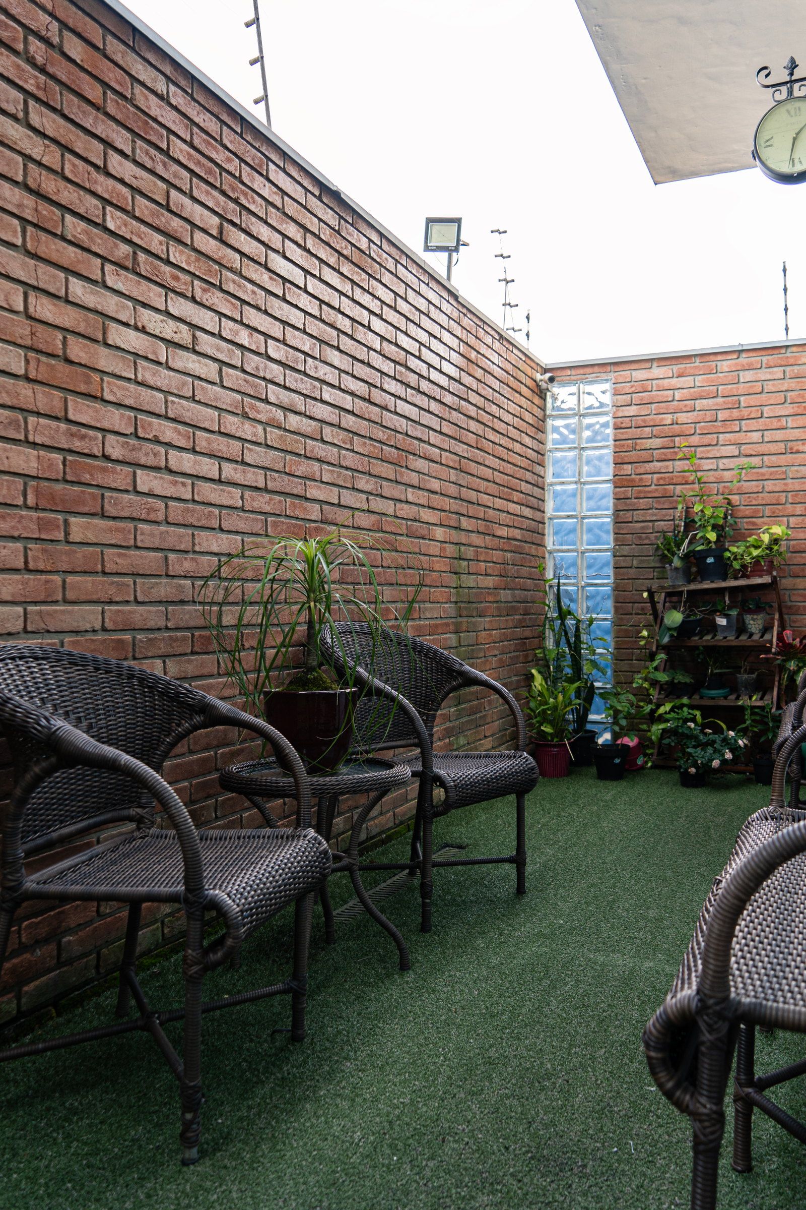 Poltronas de fibra junto ao muro de tijolos, com plantas numa estante e um relógio de parede, no Residencial Espaço Saúde