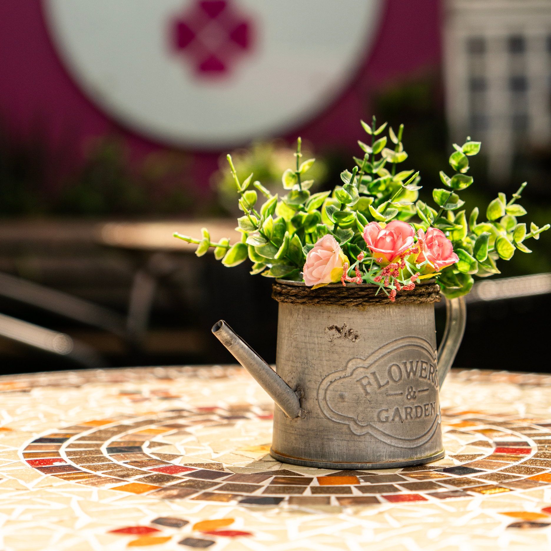 Regador com flores sobre a mesa de mosaico do jardim, com a placa da La Vie desfocada ao fundo
